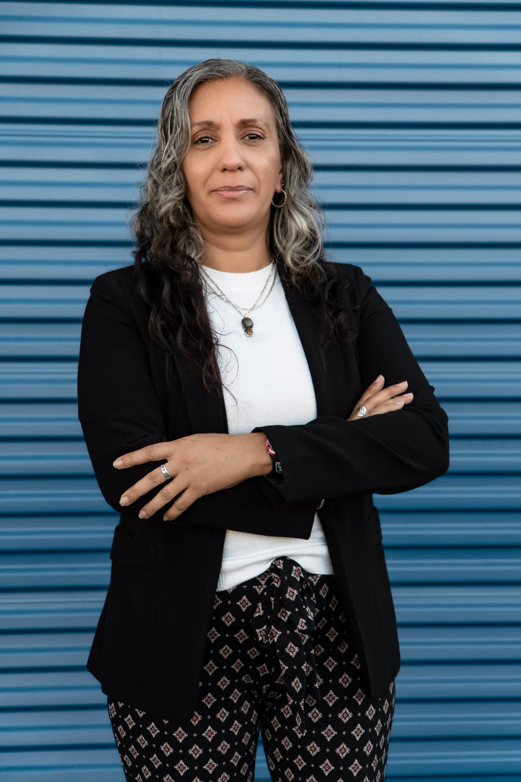 Nilsen “Nelly” Moody standing with arms crossed in a black blazer over a white top, blue background, and patterned pants, conveying confidence and professionalism