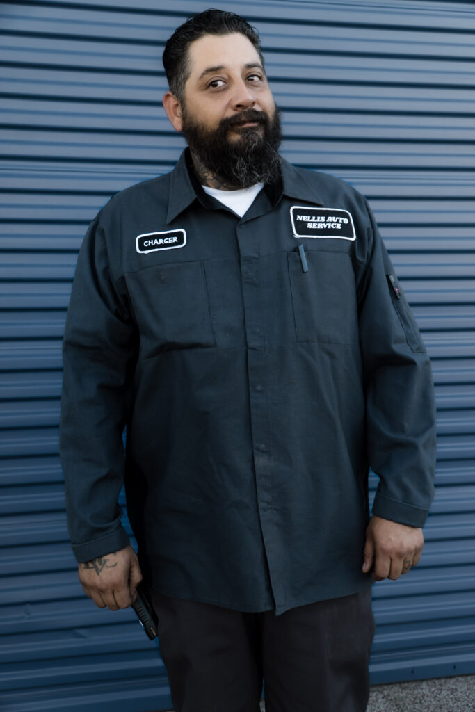 Alejandro Puente, nicknamed Charger, stands in front of a blue corrugated backdrop wearing a navy Nellis Auto Service work shirt with 'Charger' and 'Nellis Auto Service' patches, exuding a confident presence.