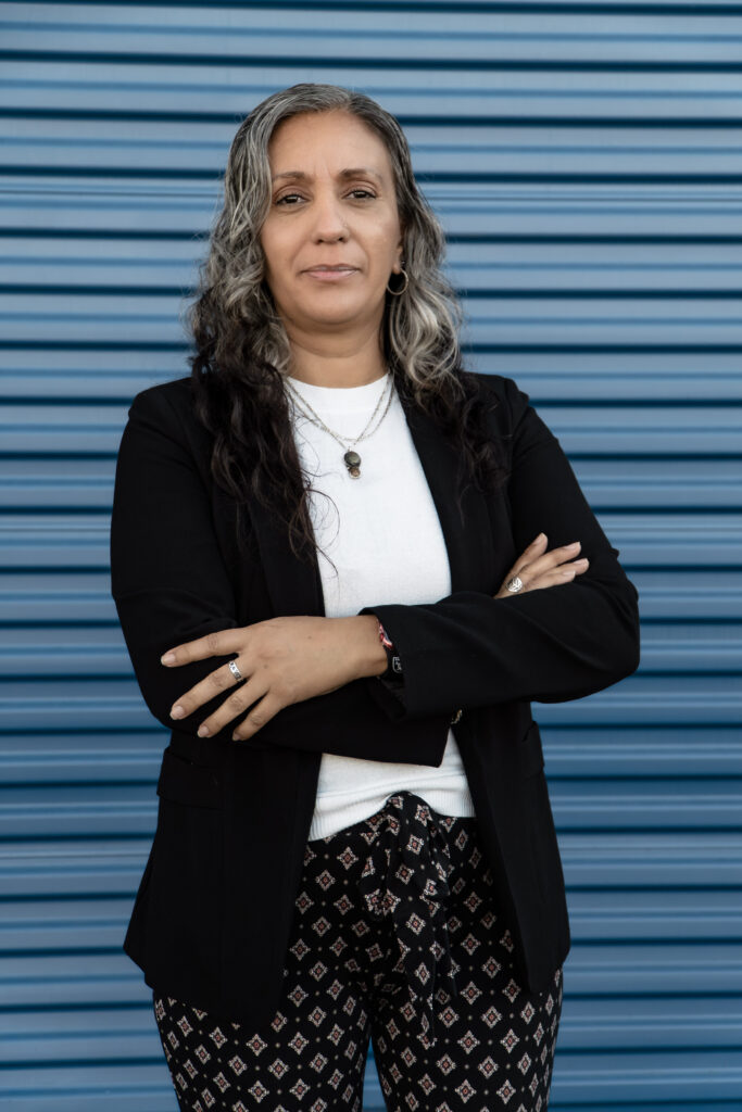 Nilsen “Nelly” Moody standing with arms crossed in a black blazer over a white top, blue background, and patterned pants, conveying confidence and professionalism